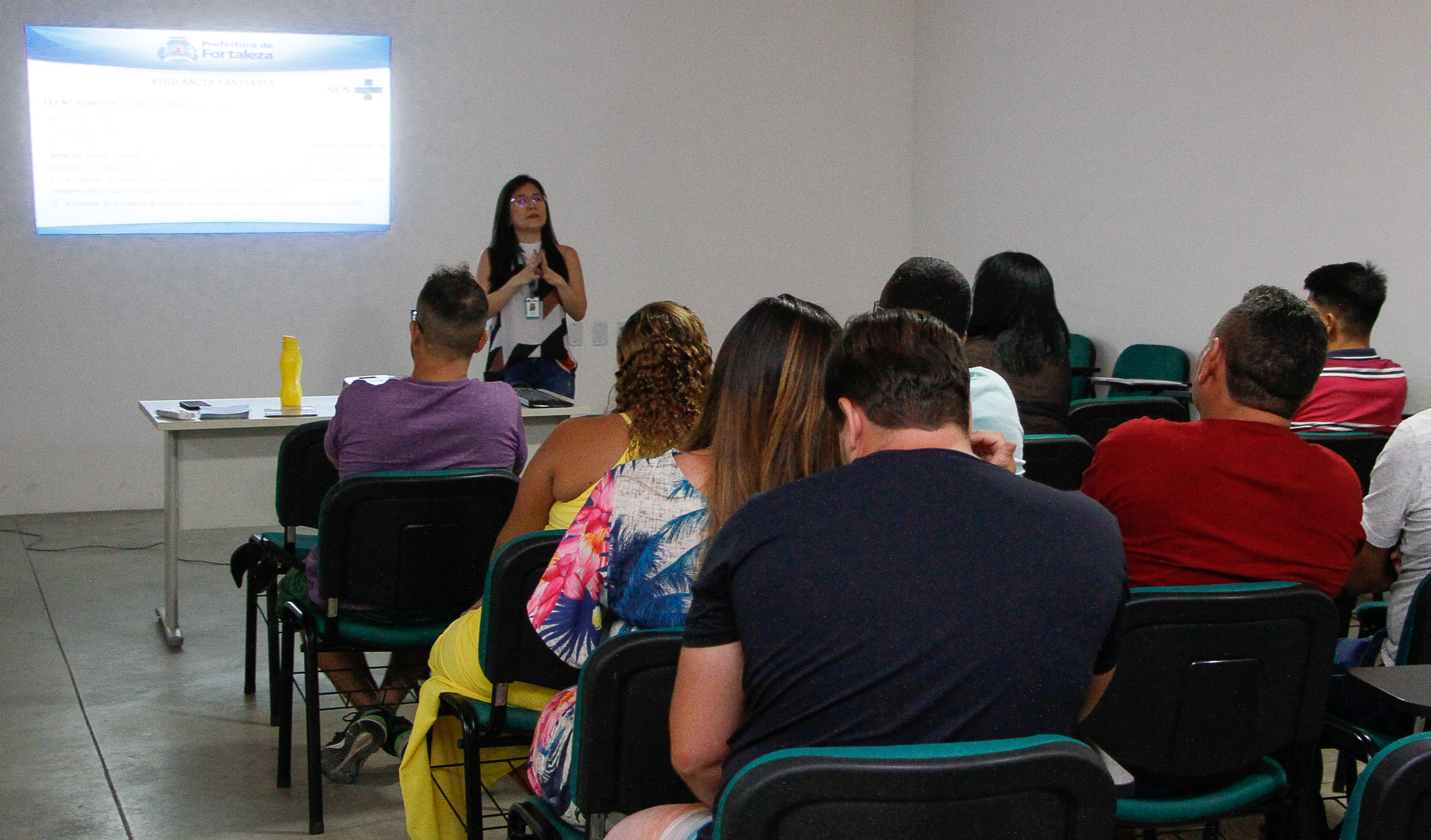 sala de aula com mulher falando à frente e pessoas sentadas assistindo